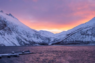 Norveç 'in Senja Adası' ndaki Torsken 'de kışın karlı dağların üzerinde gün doğumu. Karla kaplı dağ sıraları, soğuk fiyort suları ve yükselen güneşli kış manzarası. Dramatik gökyüzü manzaralı.