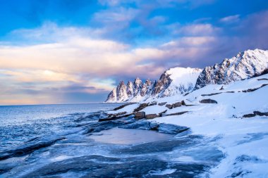 Tungeneset or Devils Jaw mountain on Senja island, Norway in winter. Winter landscape with snowy mountain peaks, fjord waters and ice coast in northern Norway. Scenic view with dramatic sky. clipart