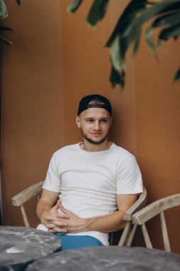 Beyaz tişörtlü ve mavi şortlu genç yakışıklı bir adam turuncu arka planda bir sandalyede oturuyor. Yakışıklı bir adamın portresi.