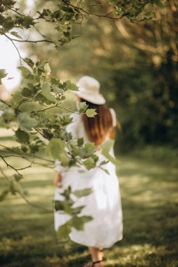Beyaz elbiseli ve şapkalı bir kadın yazın parkta yürüyor.