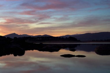 Parlak renklerle sersemletici günbatımı, kayalardan ve uzak dağlardan oluşan karanlık siluetlerle çerçevelenmiş sakin bir göle yansıyor. Huzurlu atmosfer ve doğal güzellik onu rahatlama, seyahat ve doğa temaları için mükemmel kılıyor..