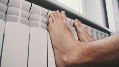 Erkek çıplak ayaklar evdeki ısıtıcı kalorifere dokunarak ısınmaya çalışır. Kış mevsiminde apartmanın ısıtıcı kaloriferlerine yakın çekim donmuş bacaklar. Isıtma sistemi zayıf, sıcaklık yok.