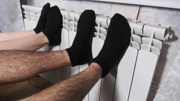 穿着袜子的男女脚在墙边的暖气散热器上加热 冻僵的情侣暖腿触摸家庭取暖电池 并试图热身 供热系统差 寒冷季节 — 图库视频影像