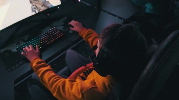 コンピュータクラブの少年は ゲームチェアでシューティングゲームをプレイ監視することによって座っています ネオンライト付きのスポーツサイバークラブでヘッドフォンのティーンエイジャーは ビデオゲームに焦点を当てています Eスポーツ バーチャルトーナメント — ストック動画