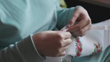 Güzel kadın elleri iplik ve iğneyle elle dikilmiş. Genç bir kadın rahat bir kanepede ev kıyafetleri içinde otururken dikiş diker. Ev atmosferi ve pencereden gelen güneş ışığı. Yaşam tarzı, el yapımı. 4K