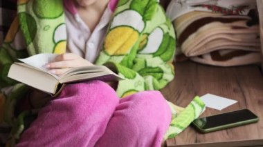 A woman reads a book sitting on a cozy sofa in daylight from the window. A female in a dressing gown is busy reading. Enjoying a book in a living room. Reading a book in a homely atmosphere. Lifestyle