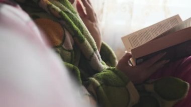 Woman is busy reading a book while sitting on a cozy sofa at home. A young woman in homely casual clothes reads in a cozy atmosphere in natural daylight from the window. Lifestyle, leisure, education