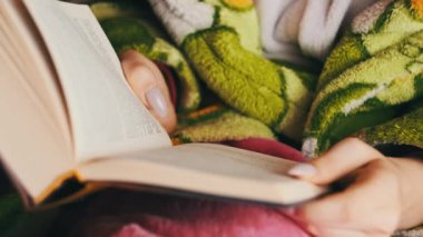 Woman is busy reading a book while sitting on a cozy sofa at home. A young woman in homely casual clothes reads in a cozy atmosphere in natural daylight from the window. Lifestyle, leisure, education