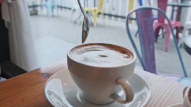 Kadın eli, caddeye bakan kahve dükkanında kaşıkla bir fincan kahvenin içinde köpük karıştırıyor. Soğuk sokağın karşısındaki sıcak kafe atmosferi. Beyaz tabakta köpüklü bir kupa kapuçino. Şehir trafiği, insanlar