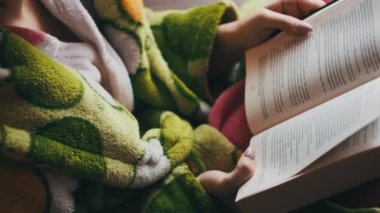 A woman reads a book sitting on a cozy sofa in daylight from the window. A female in a dressing gown is busy reading. Enjoying a book in a living room. Reading a book in a homely atmosphere. Lifestyle