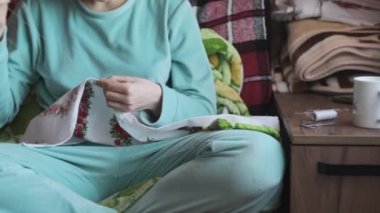 Genç kadın yavaş çekimde iplik ve iğneyle elle dikiş dikiyor. Evdeki rahat bir kanepede otururken günlük ev kıyafetleri içinde nakış yapan bir kadın. Sıcak atmosfer, doğal gün ışığı, yaşam tarzı, el yapımı.