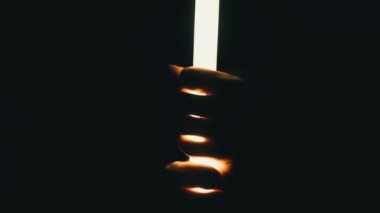Male hands hold and touch a lighting wand against a black background. Male fingers hold a bright long LED tubular lantern. Neon lightsaber. Silhouette of hands through flashlight light. 4K