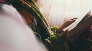 Woman is busy reading a book while sitting on a cozy sofa at home. A young woman in homely casual clothes reads in a cozy atmosphere in natural daylight from the window. Lifestyle, leisure, education