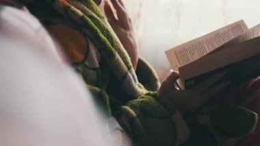 Woman is busy reading a book while sitting on a cozy sofa at home. A young woman in homely casual clothes reads in a cozy atmosphere in natural daylight from the window. Lifestyle, leisure, education