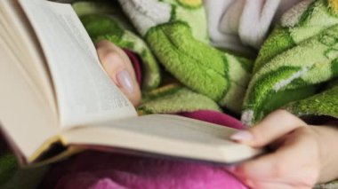 A woman reads a book sitting on a cozy sofa in daylight from the window. A female in a dressing gown is busy reading. Enjoying a book in a living room. Reading a book in a homely atmosphere. Lifestyle