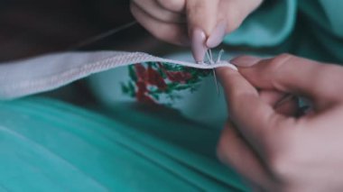 Genç kadın elleri yavaş çekimde iplik ve iğneyle elle dikilir, yakın çekimde. Manikürlü kadın parmakları evde günlük kıyafetlerle otururken dikiş dikiyor. Sıcak atmosfer, yaşam tarzı, el yapımı.