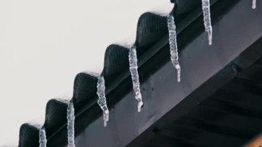 Lot of icicles hang from the overhang of the roof of the building against sky. Icicles hang from the roof of a house on a winter day. Row of icicles on the edge of house. 4K