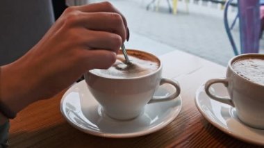 Kadın eli, caddeye bakan kahve dükkanında kaşıkla bir fincan kahvenin içinde köpük karıştırıyor. Soğuk sokağın karşısındaki sıcak kafe atmosferi. Beyaz tabakta köpüklü bir kupa kapuçino. Şehir trafiği, insanlar