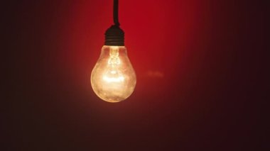 An incandescent lamp lights up when a finger touches it on dark red background. Glass bulb on wire glows, flickers, and slowly turns on and off close-up. Flashing filament. Copy space, place for text