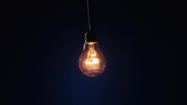 Incandescent lamp on wire glows and flickers on dark blue background. Edison bulb slowly turned on and off, pulsing light close-up. Warm flashing filament. Old classic bulb. Copy space, place for text