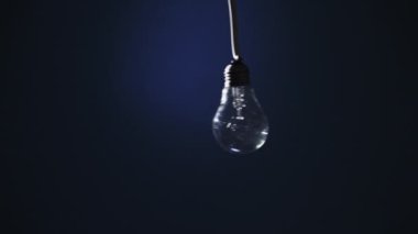 Incandescent lamp on wire sways from side to side and lights up with flickers on dark blue background. Classic bulb turned on and off close-up. Tungsten bulb cozy shine of vintage light. Copy space 4K