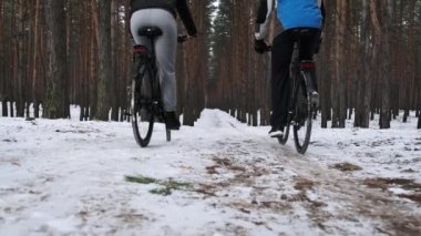 A couple of man with woman ride a bicycle along a snowy path in a winter pine forest, slow motion. Biking couple follow track between snowy trees. Two cyclists travel on black bikes. Lifestyle, sport