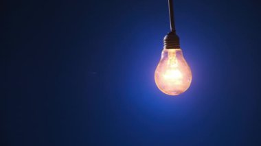 Parlayan lamba koyu mavi arka planda parlıyor ve yanıp sönüyor. Edison ampulü yavaşça açılıp kapanıyordu. Yakından yanıp sönüyordu. Ilık yanıp sönen filament. Retro klasik ampul. Boşluğu kopyala, metin için yer