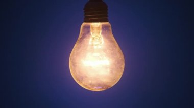 Incandescent lamp lights up and flickers on dark blue background. Classic bulb slowly turned on and off close-up. Warm flashing filament. Tungsten bulb cozy shine of vintage light. Idea, copy space 4K