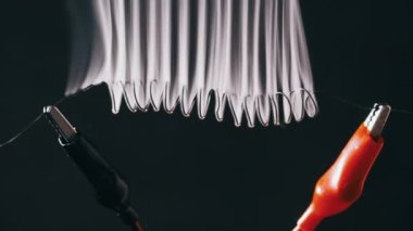 Nichrome wire heats up to red with smoke under a high electric current, slow motion. Nichrome filament in the form of spiral connected to power wires. Close-up, high metal resistance. Current strength