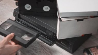 Male hand inserts VHS cassette into a VCR Video recorder. Black vintage videotape cassette recorder on a desk with many video cassettes. Inserting old VHS Tape into a retro player. Home video archive