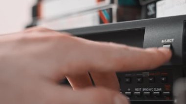 Male hand inserts VHS cassette into a VCR Video recorder. Black vintage videotape cassette recorder on a desk with many video cassettes. Inserting old VHS Tape into a retro player. Home video archive