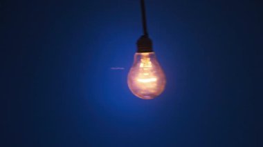 Incandescent lamp on wire sways from side to side and lights up with flickers on dark blue background. Classic bulb turned on and off close-up. Tungsten bulb cozy shine of vintage light. Copy space 4K