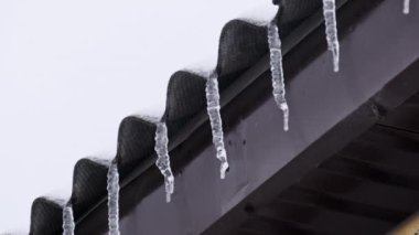 Icicles hang from the roof of a house in a winter day. Row of winter icicles hang from the overhang of the roof of the building against sky. Lot of icicles on the edge of house. 4K