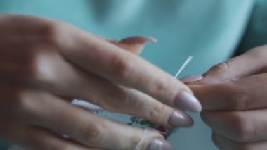 A young woman sews manually with thread and needle in slow motion, close-up. Female fingers with manicures sew while sitting in casual clothes on the sofa at home. Cozy atmosphere, lifestyle, handmade