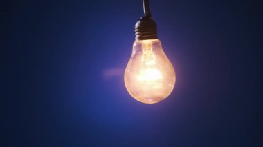 Incandescent lamp on wire sways from side to side and lights up with flickers on dark blue background. Classic bulb turned on and off close-up. Tungsten bulb cozy shine of vintage light. Copy space 4K