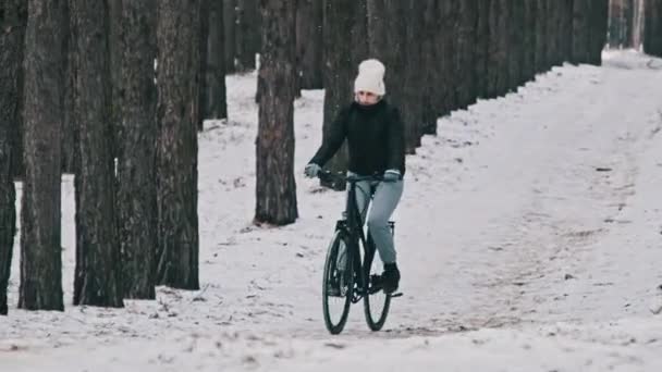 一人の女性サイクリストが風景の森の歩道に乗っています 若い女性は 松林の冬の木の間の雪の道を自転車に乗っています 健康的なライフスタイル スポーツ — ストック動画