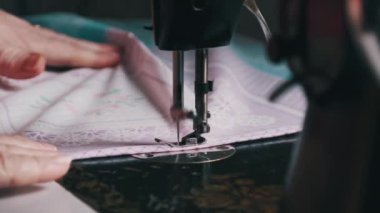 An aged seamstress sews at a traditional sewing machine at home in slow motion. Close-up of aged female hands working at a retro sewing machine. The needle quickly moves up and down. Lifestyle, hobby