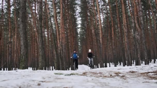 2人は一緒にゆっくりとした動きで雪の木の間の冬の道に沿って2台の自転車に乗る 女と男の自転車は田舎道をたどります 2人の自転車が黒い自転車で移動する ライフスタイル スポーツ — ストック動画