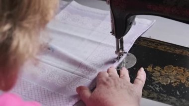 An old seamstress sews on a vintage sewing machine at home. Needle for retro sewing machine in slow motion. The process of sewing the fabric. Close-up of grandmother hands working at a sewing machine