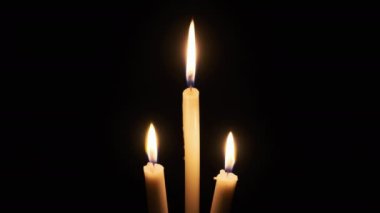 Three candle burns on a black background. Close-up of isolated yellow flickering flames illuminate the darkness. Copyscape. The warm fire of candle flame moving with the soft wind. Place for text. 4K