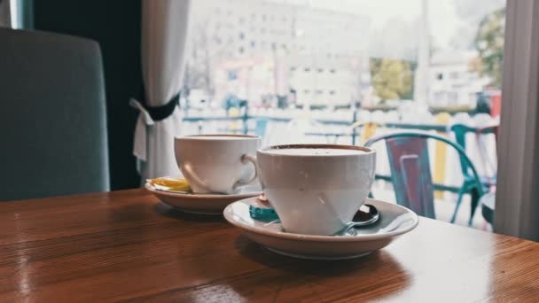 通りを見下ろす窓の反対側のコーヒーショップのテーブルの上にコーヒー 冷たい通りを見下ろす暖かいカフェの雰囲気 白い皿の上に泡とカプチーノの2つのマグカップ 都市交通 — ストック動画