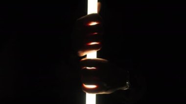 Male hands hold and touch a lighting wand against a black background. Male fingers hold a bright long LED tubular lantern. Neon lightsaber. Silhouette of hands through flashlight light. 4K