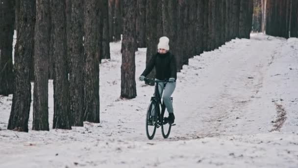 若い女性は 松林の冬の木の間の雪の道を自転車に乗って ゆっくりと動きます 風景の森の歩道で一人サイクリストが乗っています 自転車乗りは自転車に乗る 健康的なライフスタイル スポーツ — ストック動画