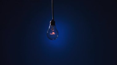 Incandescent lamp on wire sways from side to side and lights up with flickers on dark blue background. Classic bulb turned on and off close-up. Tungsten bulb cozy shine of vintage light. Copy space 4K