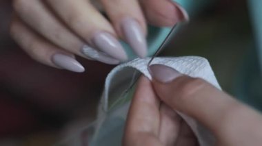 A young woman sews manually with thread and needle in slow motion, close-up. Female fingers with manicures sew while sitting in casual clothes on the sofa at home. Cozy atmosphere, lifestyle, handmade