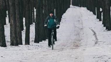 Çam ormanlarında kış ağaçları arasında karlı bir yolda bisiklet süren genç bir adam, ağır çekimde. Orman yolunda bisiklet süren bir bisikletçi var. Bisikletli bir sürücü. Sağlıklı yaşam tarzı, aktif hobiler, spor