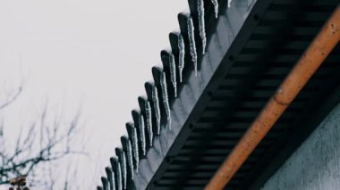 Lot of icicles hang from the overhang of the roof of the building against sky. Icicles hang from the roof of a house on a winter day. Row of icicles on the edge of house. 4K