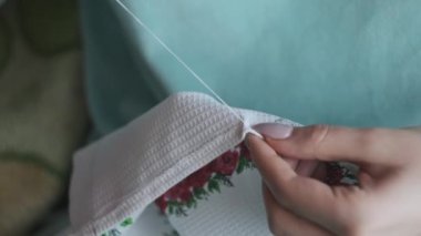 Güzel kadın elleri iplik ve iğneyle elle dikilmiş. Genç bir kadın rahat bir kanepede ev kıyafetleri içinde otururken dikiş diker. Ev atmosferi ve pencereden gelen güneş ışığı. Yaşam tarzı, el yapımı. 4K