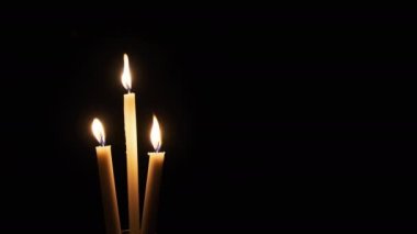 Three candle burns on a black background. Close-up of isolated yellow flickering flames illuminate the darkness. Copyscape. The warm fire of candle flame moving with the soft wind. Place for text. 4K