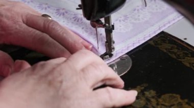 Old female hands of seamstress sew at a retro sewing machine at home in slow motion. Close-up of aged female fingers sewing fabric at a traditional sewing machine. The needle quickly moves up and down